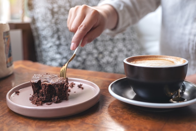 カフェの木製テーブルにコーヒーカップとフォークでブラウニーケーキを切る女性