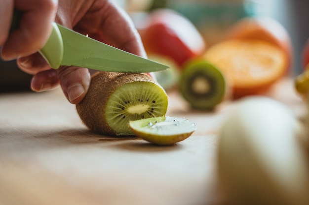 A woman cutting a kiwifruit with a green knife on a wooden table surrounded by other fruits in the morning.