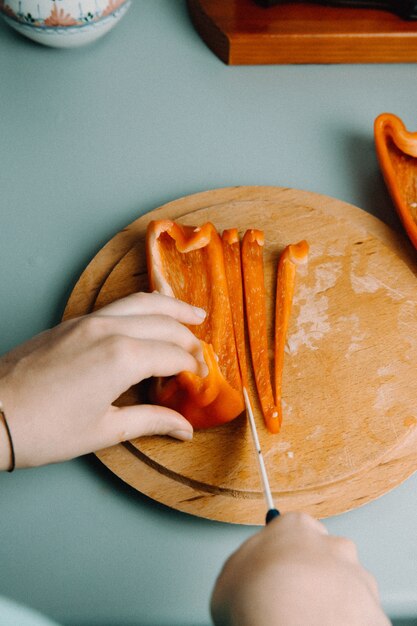 A woman cutting some pepper over a wooden round plate with a knife while cooking with the pan in the kitchen