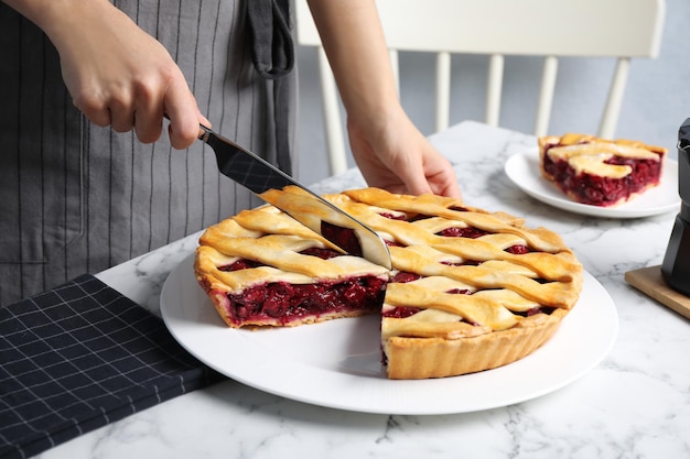 Woman cutting tasty cherry pie at white marble table closeup