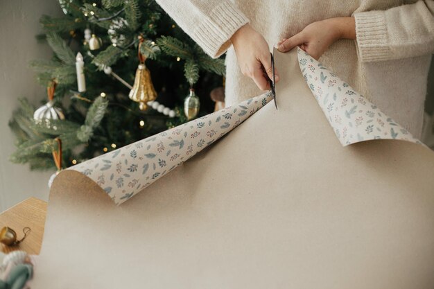 여성이 가위로 절단하는 스타일리시 크리스마스 선물을 포장하기위한 축제 종이 현대적으로 장식 된 스칸디나비아 방에서 근접하여 겨울 휴가 준비