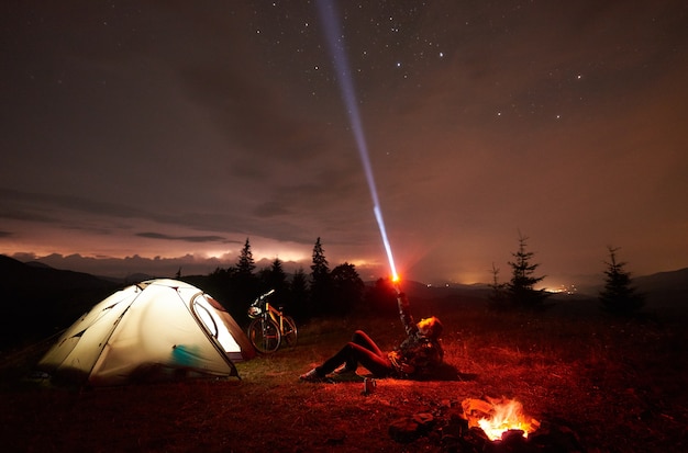 Woman cyclist at night camping near burning campfire