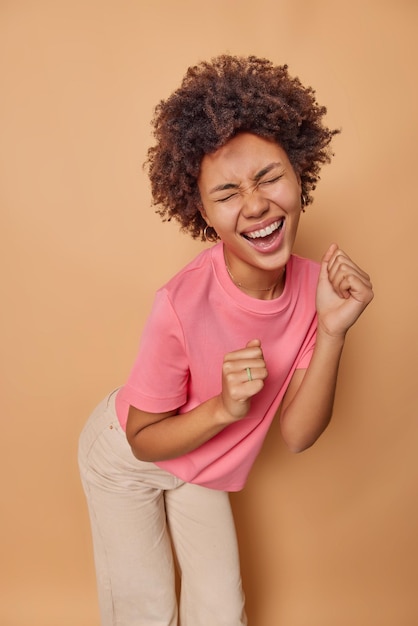 Photo woman dances and shakes fists feels upbeat and energetic wears casual pink t shirt trousers closes eyes from happiness isolated on beige. happiness and joy concept