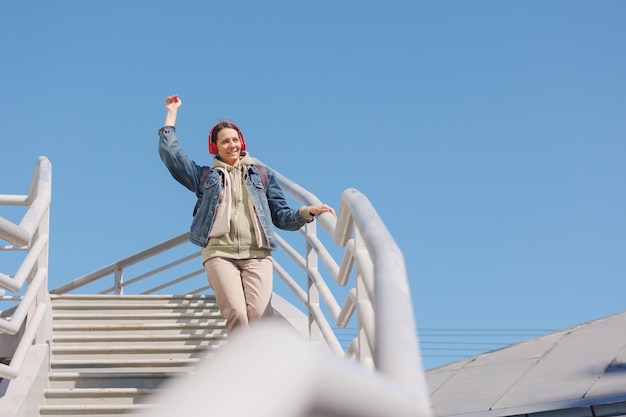 Woman dances and sings on stairs listens to music or podcast with red headphones fulllength