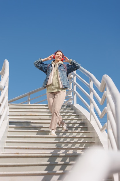 Woman dances and sings on stairs listens to music or podcast with red headphones fulllength