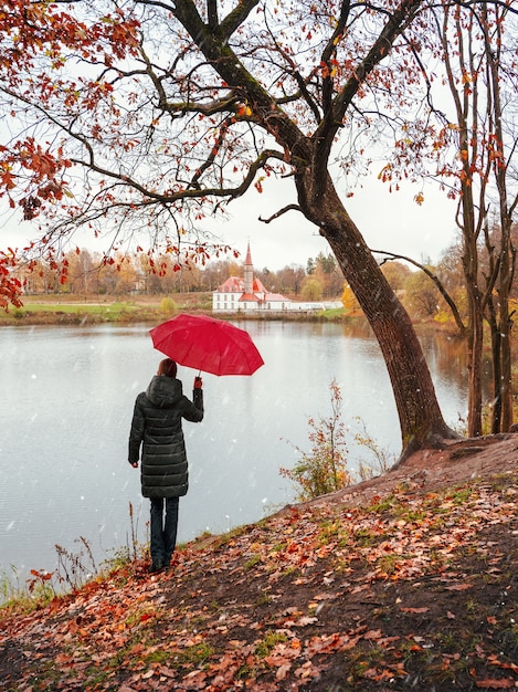 Woman in a dark coat and a red umbrella looks into the distance in a snowy and rainy autumn.