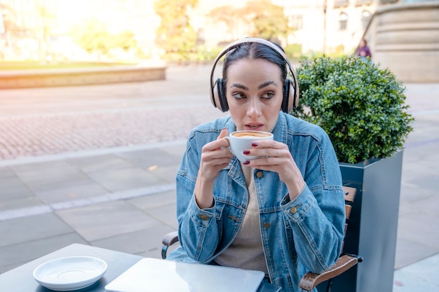 Женщина в джинсовом пальто и наушниках пьет кофе и слушает музыку Концепция образа жизни