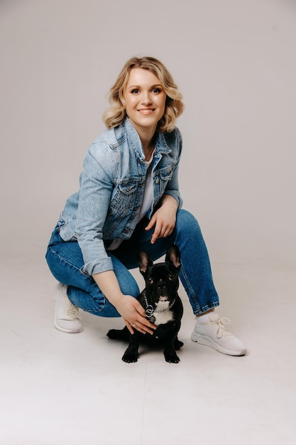 A woman in a denim jacket and jeans poses with a french bulldog.