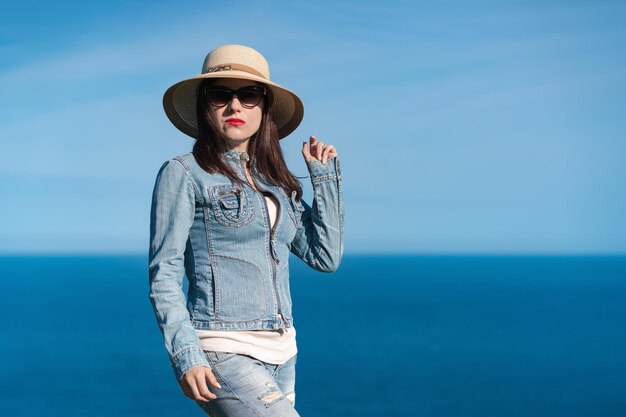 Woman in denim jacket jeans straw hat and sunglasses standing against backdrop of blue sky and sea