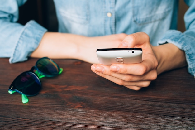 Foto la donna in una camicia del denim tiene un telefono cellulare e sedendosi ad una tavola di legno su cui si trovano gli occhiali da sole