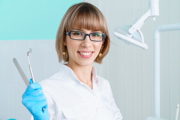 Woman dentist holding tools on background of the dental office in a white coat and glasses smiles th...