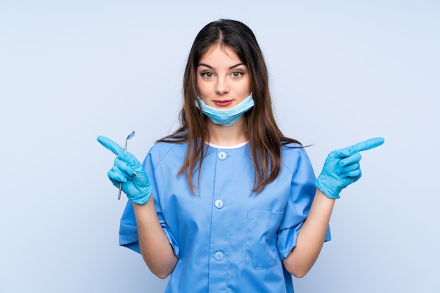 의심을 가지고 측면을 가리키는 파란색 벽 위에 도구를 들고 여자 치과 의사