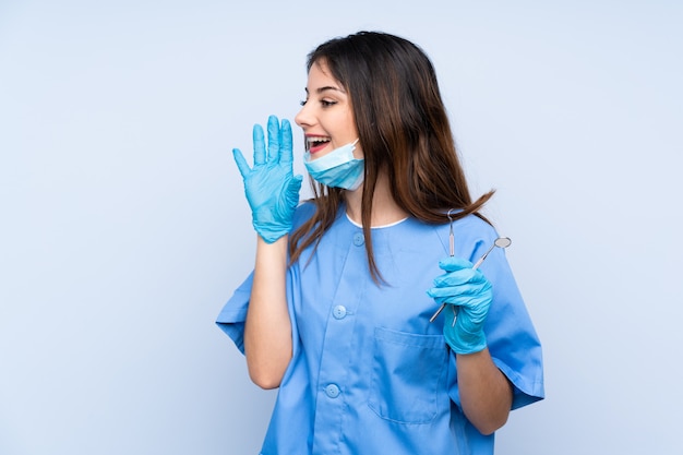 Woman dentist holding tools over blue wall shouting with mouth wide open