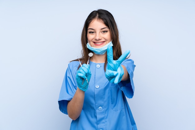 Woman dentist holding tools over blue wall smiling and showing victory sign