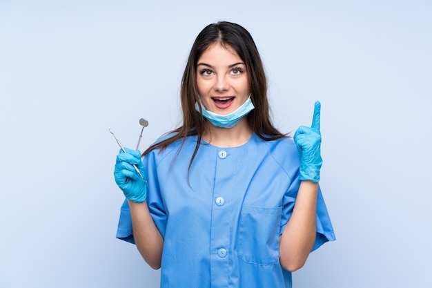 Woman dentist holding tools over isolated blue wall pointing with the index finger a great idea