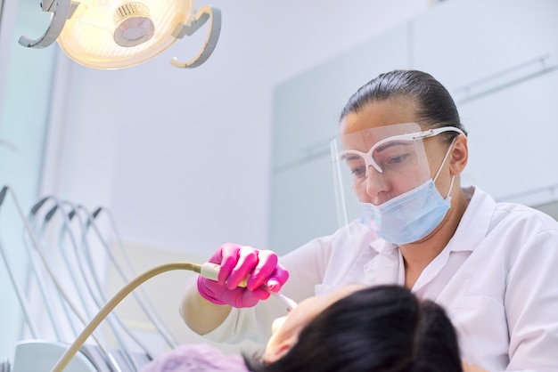 Woman dentist treating teeth to a patient sitting in dental chair using professional equipment