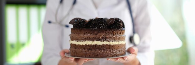 女性栄養士はクリームとチョコレート ケーキの大きなスライスを保持している医者は食べないようにアドバイスします