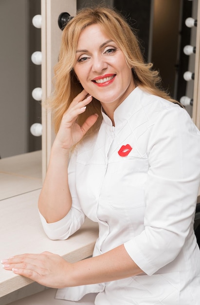 A woman doctor beautician poses against the background of mirrors with light bulbs, looks into the camera and smiles. Beauty and self care concept