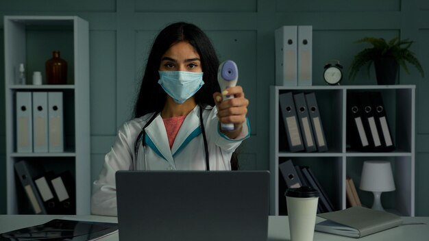 woman doctor in mask measuring temperature with digital scanner in hospital room on laptop webcam