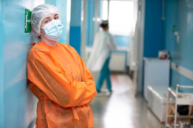 Woman doctor in a medical mask protective gown and hat looks at the camera Tired female surgeon