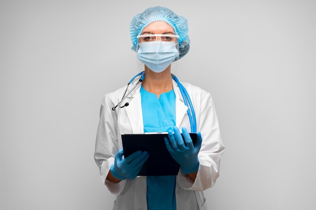 Woman doctor in medical mask standing with clipboard against gray background, portrait