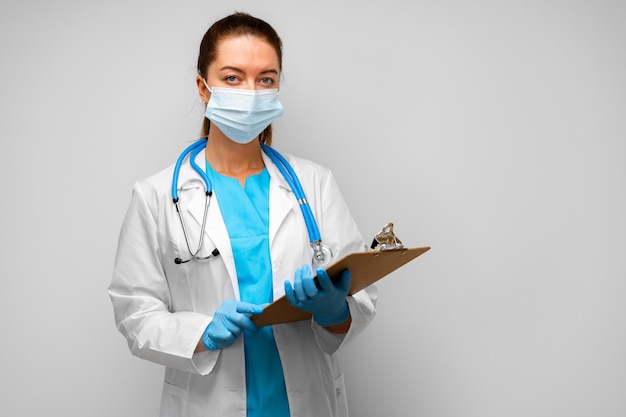 Woman doctor in medical mask standing with clipboard against gray