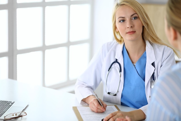 Woman doctor and patient sitting and talking at medical examination in hospital or clinic