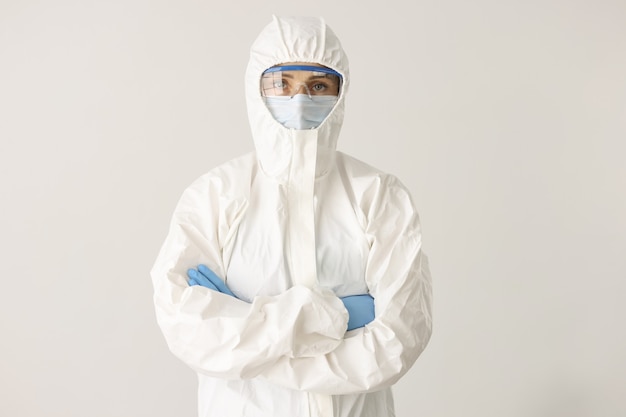 Woman doctor or scientist stands in protective suit mask and glasses with folded arms on white