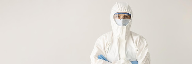 Woman doctor or scientist stands in protective suit mask and glasses with folded arms on white