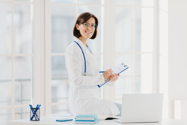 Woman doctor sits on table, writes down information in clipboard