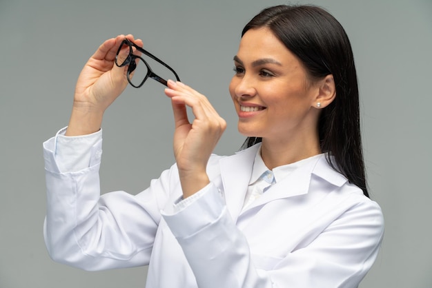 Woman doctor smiling while holding glasses and looking at it Portrait of beautiful young female doctor in white medical jacket isolated on grey background