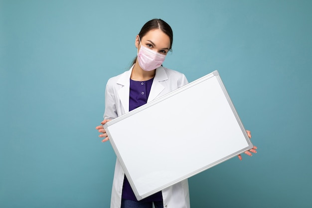 Woman doctor wearing a white medical coat and a mask holding blank board with copy space for text