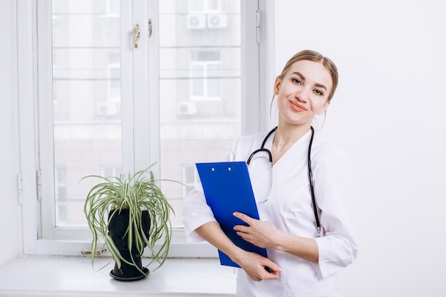 a woman doctor in a white coat with a stethoscope on her shoulders holds a blue folder and smiles by a window