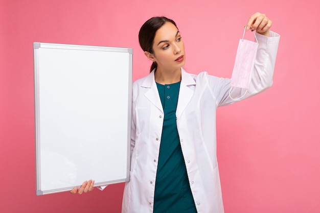 Foto medico della donna in un camice medico bianco che tiene scheda in bianco con lo spazio della copia per testo e maschera protettiva isolata su priorità bassa. concetto covid.