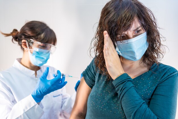 A woman doctor with a face mask applying the coronavirus vaccine, the patient with fear of the vaccine and its side effects.