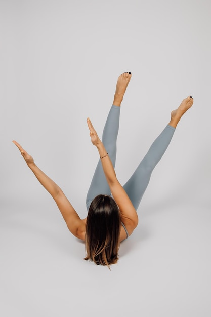 Woman does sports Young brunette performs exercise from yoga or fitness she lies on her back and lifts up her arms and legs and turns them to side gray isolated background