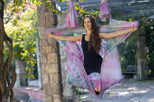 A woman doing aerial yoga  takes a pose in the hammock and smiling to the camera