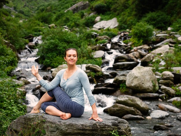 Woman doing Ardha matsyendrasana asana half spinal twist pose at tropical waterfall in Himalayas