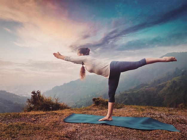 Woman doing Ashtanga Vinyasa Yoga asana Virabhadrasana 3 Warrior pose in mountains on sunset