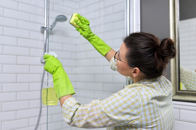 Женщина делает уборку в ванной, дома. Женское мытье душевых стекол с губкой и моющим средством.