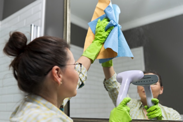 Woman doing cleaning in bathroom using vacuum cleaner, without use of household chemicals. Close-up of gloved hands with steam in the mirror, eco-friendly cleaning