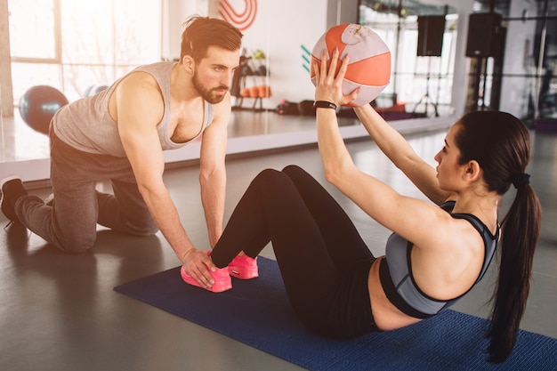 彼女のスポーツパートナーが彼女の足を床に押し下げている間にボールでいくつかの腹筋運動をしている女性。彼は彼女が適切な方法で運動をするのを助けます。