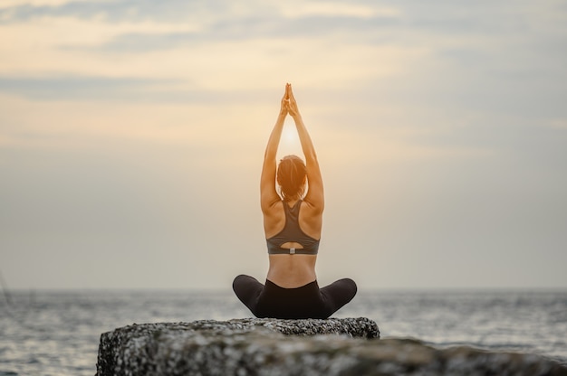 A woman doing yoga asana sitting on the stone at the sea.Morning natural stretch warm-up training.Yoga,fitness and healthy lifestyle concept.