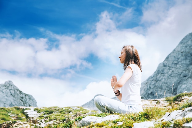 Woman doing yoga and meditating in lotus position on the background of sky and mountains