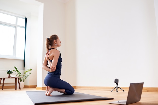 Woman doing yoga online. Computer and camera in her living room. Healthy lifestyle and distance work concepts