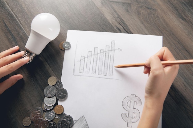 A woman draws a graph on paper and on the table there are coins and a light bulb