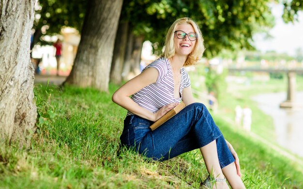 Woman dreaming about vacation Literature for summer vacation Summer vacation Student smart nerd sit on green grass and read book Girl relaxing at riverside after working day Rest relax and hobby