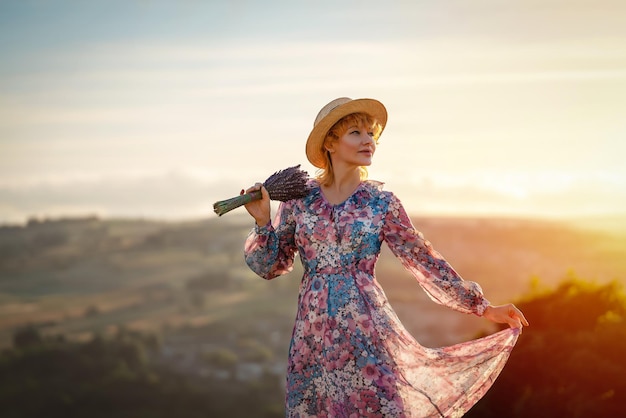 Woman in a dress and a hat with a lavender bouquet in her hands on a hill in summer sunset