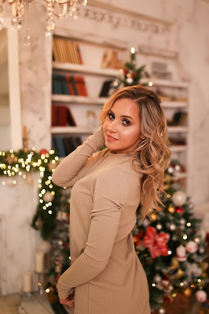 A woman in a dress posing by a tree and a fireplace in a bright interior at home in the evening