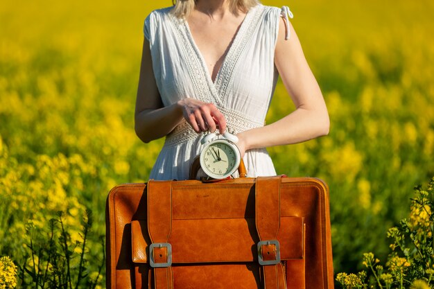 Woman in dress with suitcase and alarm clock in rapeseed field